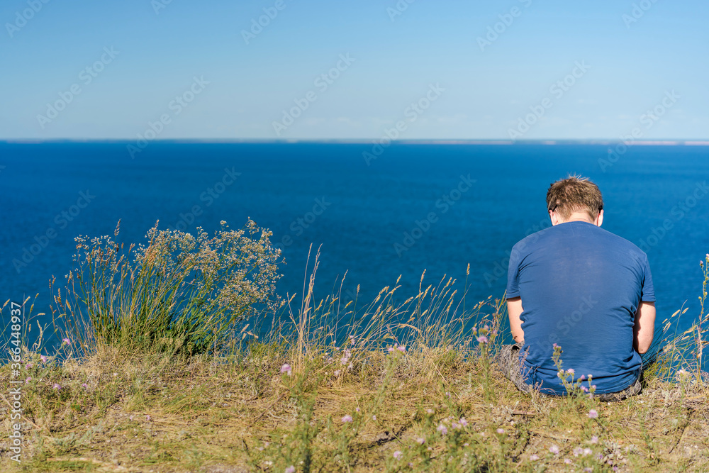 A blond man in a blue shirt sits on a cliff overlooking the blue sea and Islands