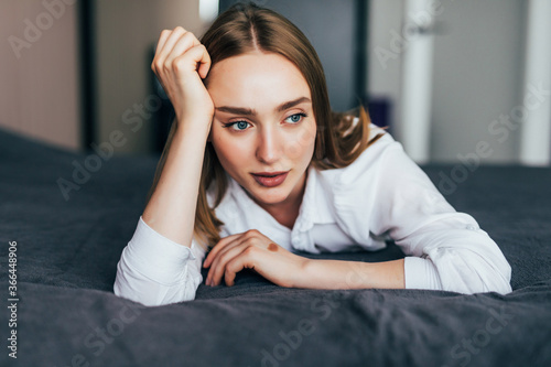 Thoughtful casual woman in white pajamas lying on her bed in a bright bedroom