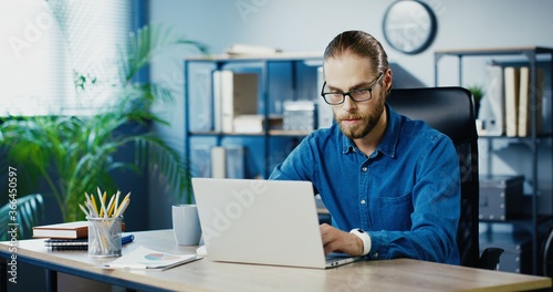 Caucasian serious male office employee typing on laptop while working in cabinet. Handsome busy man in glasses browsing on computer while sitting at workplace indoor. Business concept