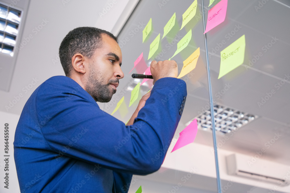 Fototapeta premium African American office employer writing on sticker with marker. Focused confident businessman in suit sharing idea for project and making note. Brainstorming, business and management concept
