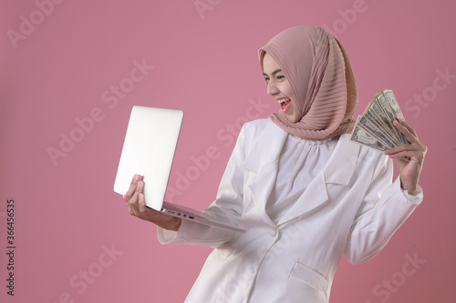 Smiling young muslim woman in hijab white clothes hold laptop pc and money, isolated on pink wall background, studio portrait. People religious lifestyle concept. Mock up copy space
