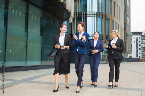 Group of female coworkers walking with takeaway coffee outdoors, talking, smiling. Full length, front view. Work break concept