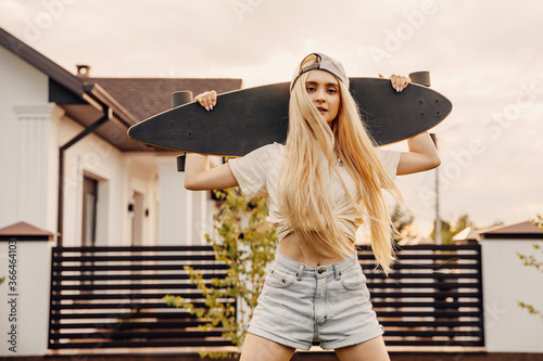 Teenage girl with long blonde hair holding a skateboard on her shoulders.