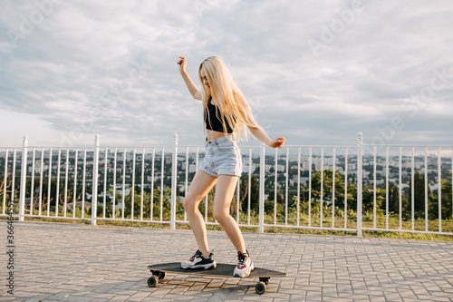 Teenage girl wearing denim shorts and black crop top, skating on a long board, in a park at the sunset.