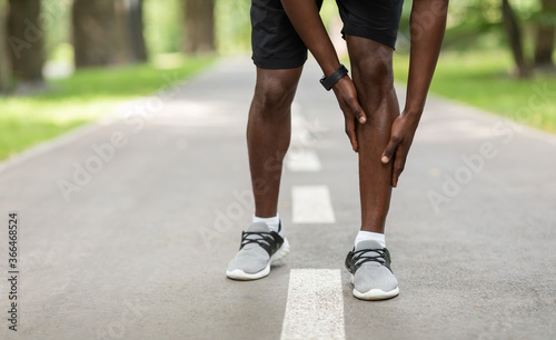 Black sportsman touching his aching leg while jogging outdoors