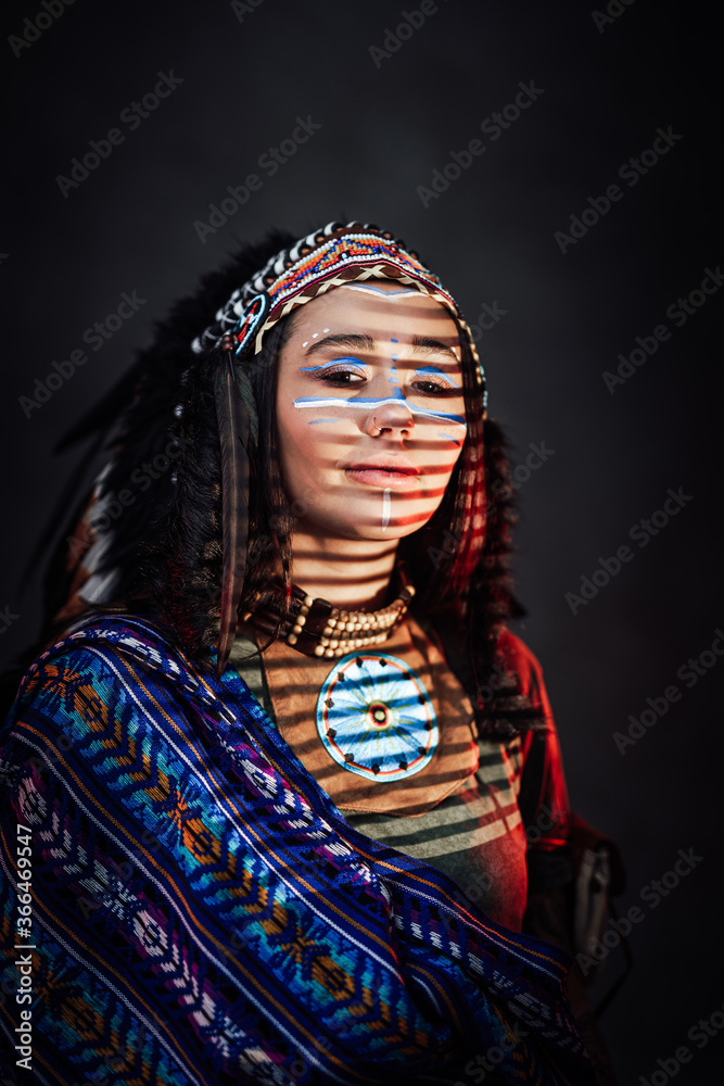 Beautiful American Indian girl with traditional make up in Indian feather hat and ethnical clothes. Studio portrait with a striped shadow from the blinds on a dark background