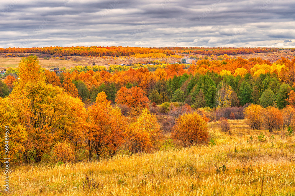 Picturesque autumn landscape in green and yellow colors. Panoramic view from hill to lowland with grove, village and field in cloudy day. Colorful autumnal nature, beautiful natural background