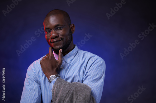African American student in blue shirt and glasses, holds hand on chin and smiling. Studio portrait on a dark background with blue light