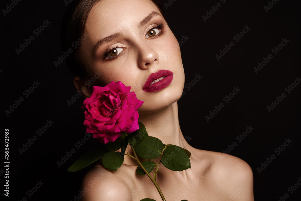 Beautiful young woman with modern make-up red lips big eyelashes holding a pink rose in her hands on a black background