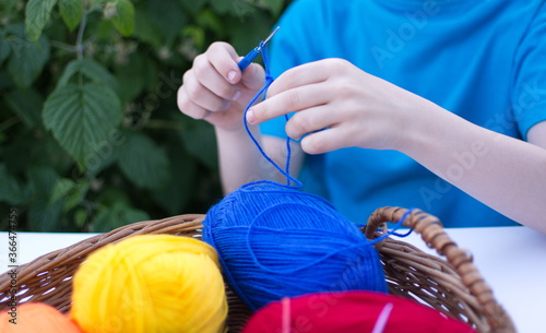 a child in a blue t-shirt is crocheting, and in a wooden basket, against the background of green leaves, there are colored skeins of woolen thread for knitting,