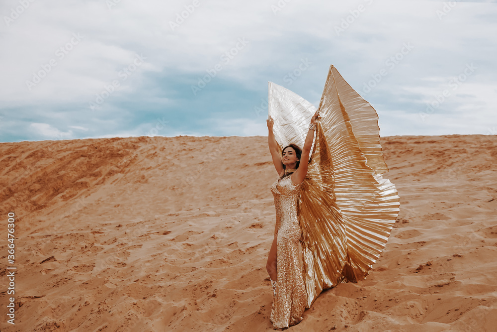 A beautiful girl in a golden with brilliant dress with wings, suit is dancing an oriental, East dance in the desert.