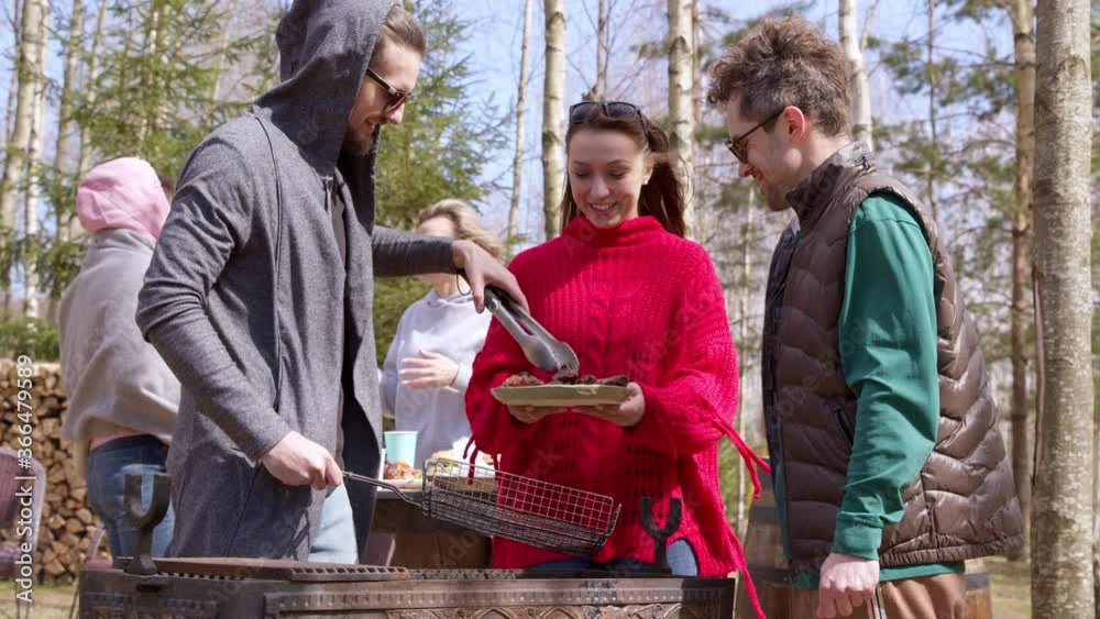Young happy people having fun camping enjoying bbq party outdoors, friends making barbecue on summer picnic, handsome guy puts grilled meat from grill lattice to plate. Food, resting time concept Young happy people having fun camping enjoying bbq party outdoors, friends making barbecue on summer picnic, handsome guy puts grilled meat from grill lattice to plate. Food, resting time concept