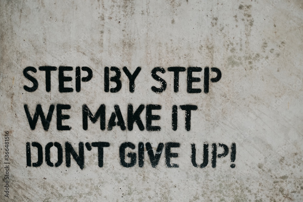 Plakat Motywacyjny cytat „krok po kroku robimy, nie poddawaj się” napisany na białej ścianie czarną farbą