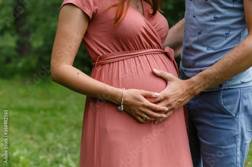 Husband hugs a pregnant woman in a pink dress. Father and mother hands on pregnant belly