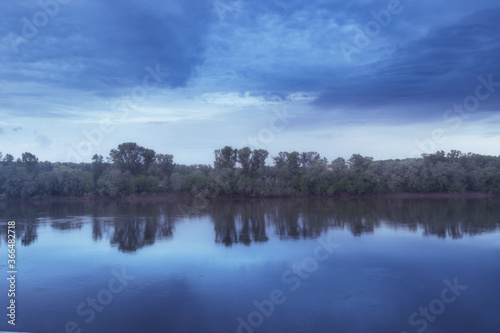 An idyllic landscape on a river or lake at dusk during the blue hour. Reflections in the water of old trees