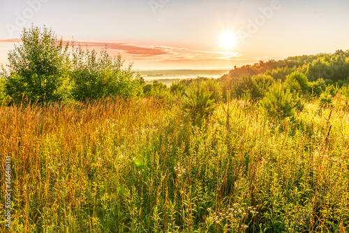 Scenic view at beautiful sunrise in a far misty valley  bright crimson cloudy sky   trees and golden sun rays with glow  summer morning  field landscape