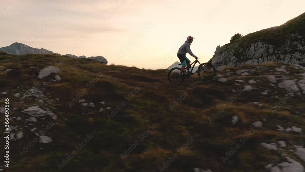 Aerial - Active young man riding the bicycle down the mountain terrain at sunset. 4K