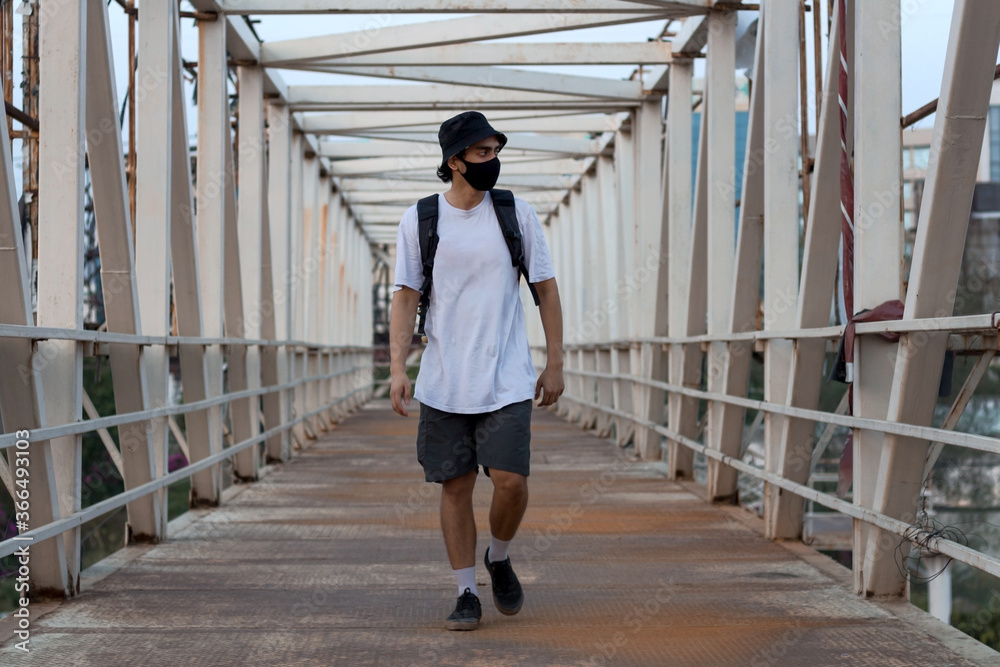 Young Millennial Walking on an Empty Metal Foot Bridge Outdoors while Wearing a Black Protective Face Mask to Prevent Covid-19 Virus Infection in a City. The New Normal.
