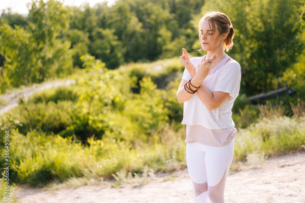 Calm young meditating young woman standing on nature with closed eyes holding crossed hands on her chest in yoga posture mudra. Concept of practicing yoga alone outdoor. Calm young meditating young woman standing on nature with closed eyes holding crossed hands on her chest in yoga posture mudra. Concept of practicing yoga alone outdoor.