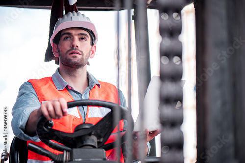 Young confident Caucasian man engineer driving Forklift and wearing white safety helmet and check for control loading containers box from Cargo freight ship for import and export, transport