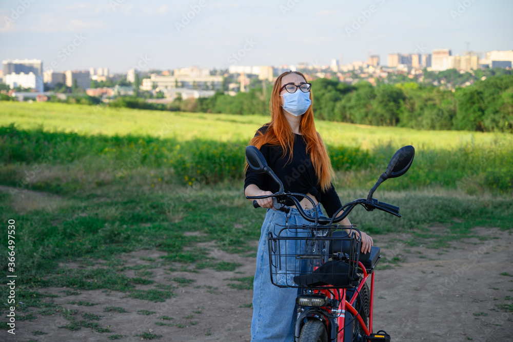 caucasian girl stand near red electro bicycle at background of blue sky puts on medical mask, looks in the mirror. lifestyle, coronavirus, health protection, quarantine, safety and pandemic concept