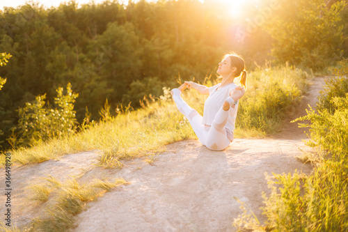 Young fit woman performing yoga posture Boat with Toe Hold, outside evening on background of bright sunray. Female practicing yoga exercises in alone outdoors during sunset.  photo