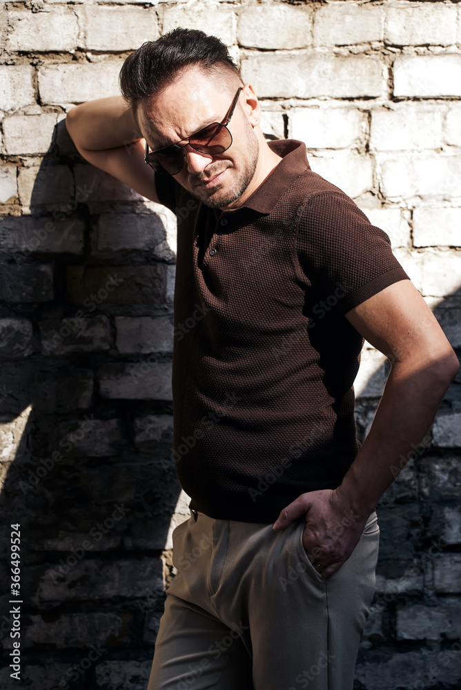 stylish guy with glasses stands near a stone wall and looks away. thought. strong and strict male gaze. brown T-shirt with collar and fashionable beige trousers. sunlight. stylish guy with glasses stands near a stone wall and looks away. thought. strong and strict male gaze. brown T-shirt with collar and fashionable beige trousers. sunlight.