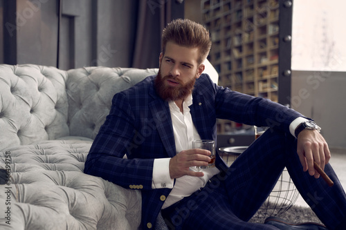 Handsome stylish man blue suit in a cage at home sitting on the floor with glass of whiskey in hand