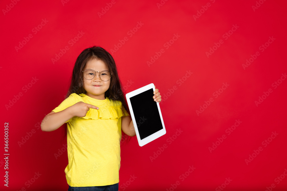 Showing blank tablet's screen. Beautiful little girl on red background. Half-lenght portrait of happy child gesturing. Cute asian girl in yellow. Concept of facial expression, human emotions Showing blank tablet's screen. Beautiful little girl on red background. Half-lenght portrait of happy child gesturing. Cute asian girl in yellow. Concept of facial expression, human emotions