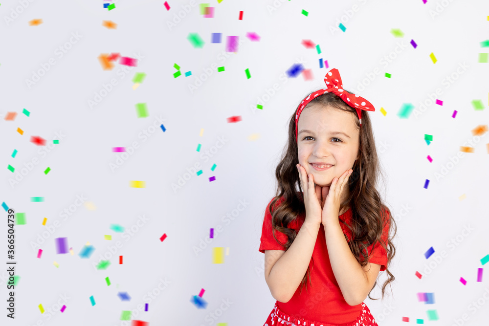 A cute little girl in a red dress on a white isolated background with a streamer folded her hands on her cheeks. Space for text. Little girl celebrates birthday, holiday concept