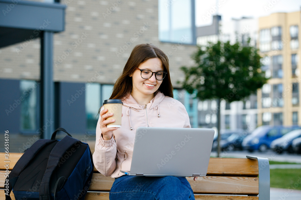 Positive girl in jeans with coffee cup in hand sits on a bench and working at a laptop. Freelance work concept