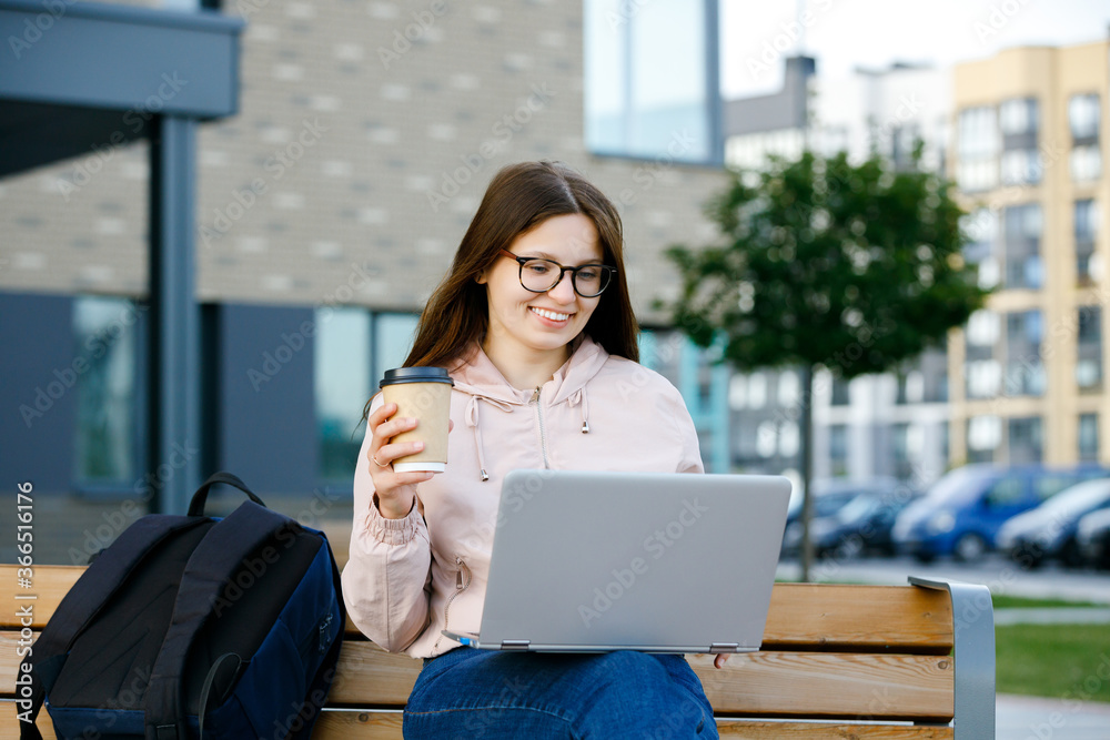 Positive girl in jeans with coffee cup in hand sits on a bench and working at a laptop. Freelance work concept Positive girl in jeans with coffee cup in hand sits on a bench and working at a laptop. Freelance work concept