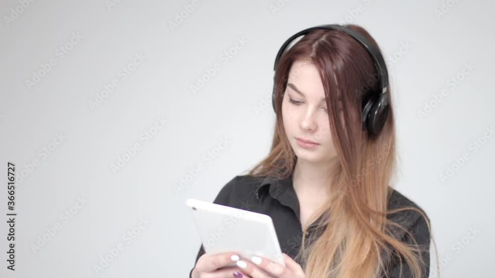 Pretty young serious good looking woman with long hair in headphones uses white tablet on white background Pretty young serious good looking woman with long hair in headphones uses white tablet on white background
