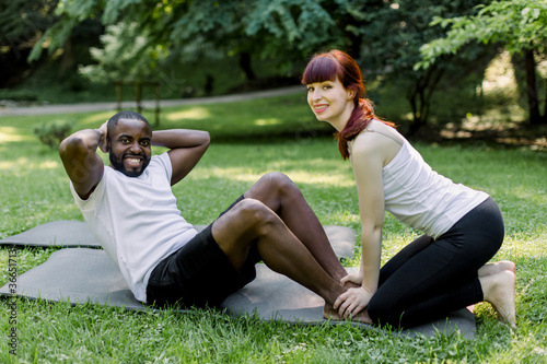 Fitness, sport, training, teamwork and people concept. Handsome muscular African man, doing sit ups in green summer park with his personal trainer, charming young Caucasian woman