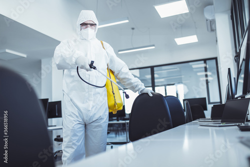 Office disinfection during COVID-19 pandemic. Man in protective suit and face mask spraying for disinfection in the office photo