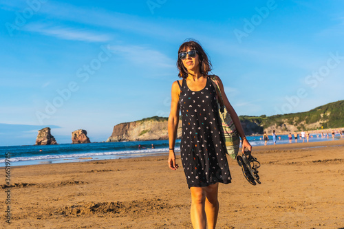 A young brunette in a black dress and sunglasses walking along the beach in Hendaye, French Basque country. France