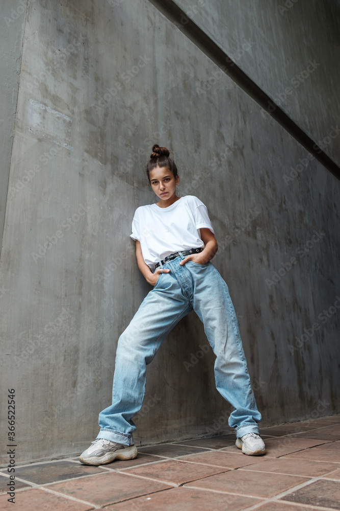 A beautiful young woman in full growth stands against the wall. A stylish girl in blue jeans and a white T-shirt stands against the background of a gray concrete wall. 80's 90's style A beautiful young woman in full growth stands against the wall. A stylish girl in blue jeans and a white T-shirt stands against the background of a gray concrete wall. 80's 90's style
