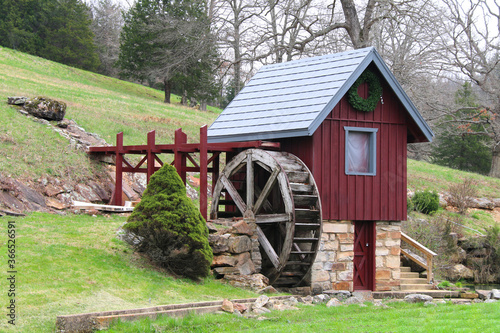 A small replica red and blue old mill shed building with wooded water wheel and stone foundation