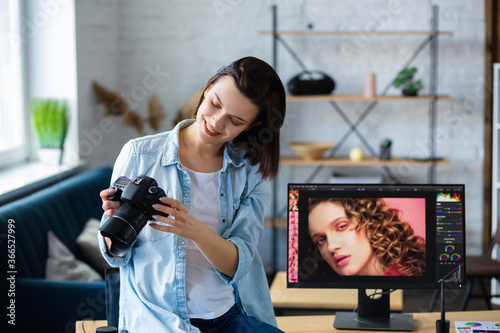 Portrait of professional photographer holding camera in photo studio. Workplace in office with camera, laptop, monitor, graphic drawing tablet and color palette. Retouching images. Creative agency.