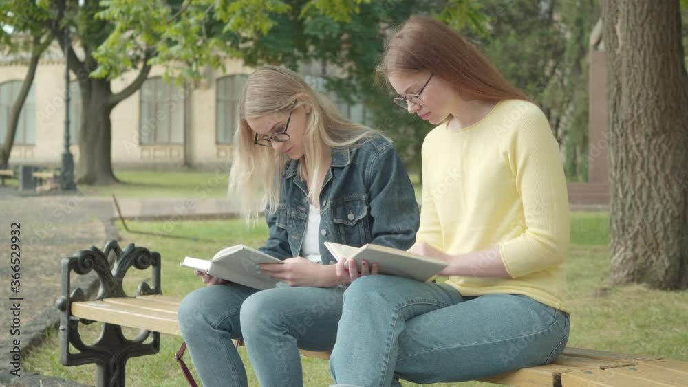 Two concentrated nerd female students in eyeglasses sitting on bench and reading books. Portrait of focused young Caucasian girls studying outdoors on university campus yard. Education, intelligence. Two concentrated nerd female students in eyeglasses sitting on bench and reading books. Portrait of focused young Caucasian girls studying outdoors on university campus yard. Education, intelligence.