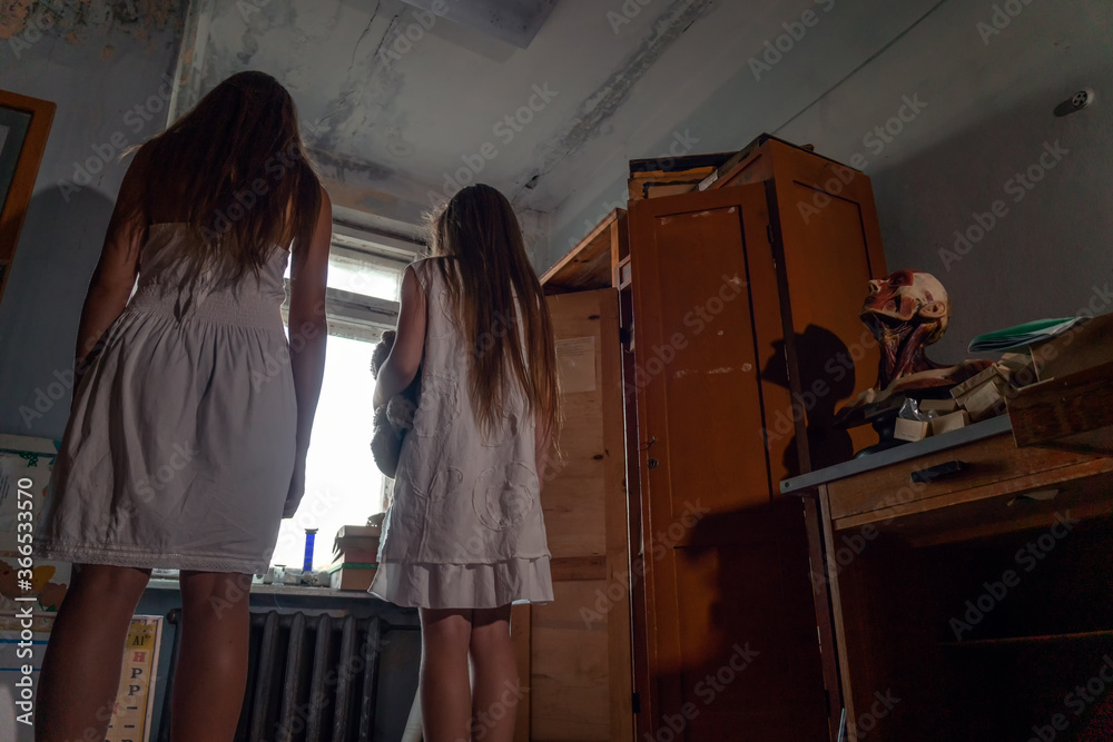 two girls in white dresses with long hair and a Teddy bear in their hands stand with their backs to the camera in a dark abandoned room with scattered belongings. concept of horror, mysticism