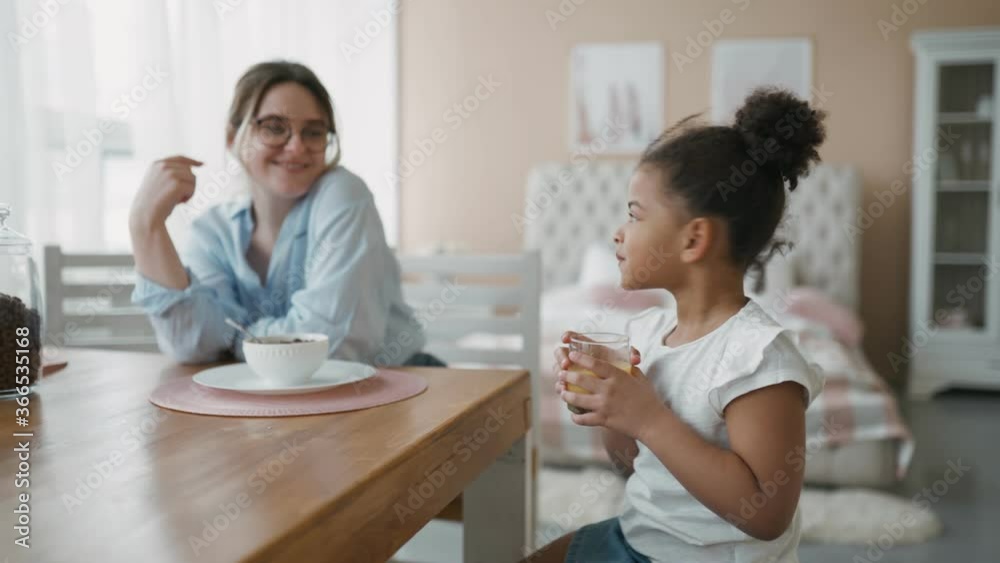 Breakfast in kitchen at home. Young European mother feeds cute, beautiful African daughter. Child gladly drinks orange juice sitting in chair in front of school. Family is having fun together concept. Breakfast in kitchen at home. Young European mother feeds cute, beautiful African daughter. Child gladly drinks orange juice sitting in chair in front of school. Family is having fun together concept.