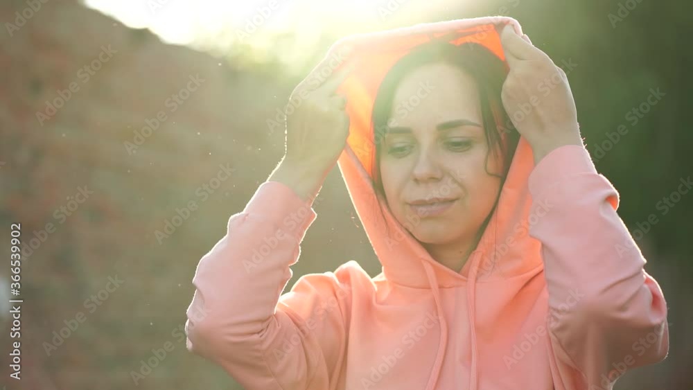 Portrait of young woman putting on hood on street. Adult female puts