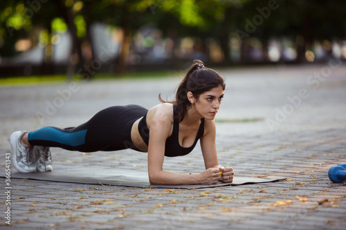 Young female athlete training in the city street in summer sunshine. Beautiful woman practicing, working out. Concept of sport, healthy lifestyle, movement, activity. Stretching, sit-ups, ABS's.