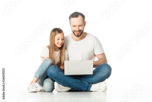 smiling father and daughter sitting on floor with laptop isolated on white