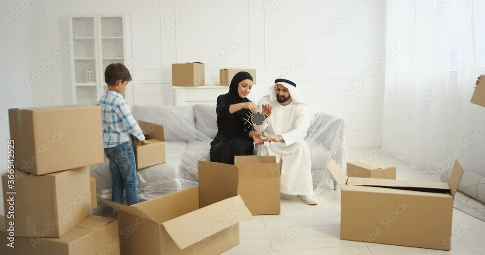 Smiled Arabic mother and father sitting on sofa in living room with small son. Unpacking carton boxes. Muslims with kid moving in new home Adults and cute child from Middle East changing accommodation