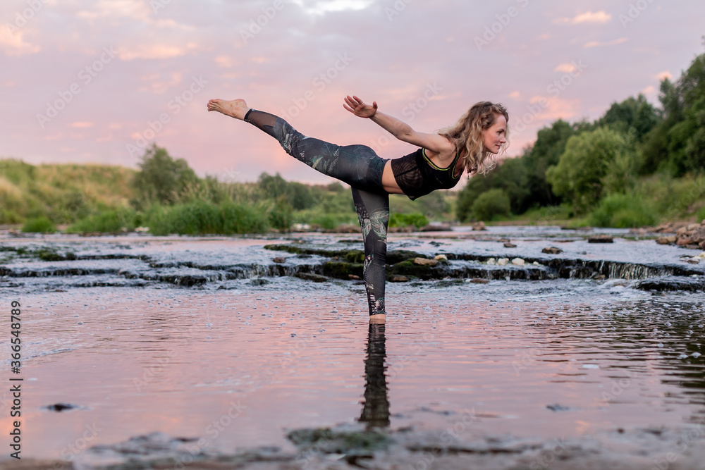 Blonde woman in sportswear practicing yoga standing in the middle of river in front of a sunset sky. Fitness girl doing a balance exercise over the water.