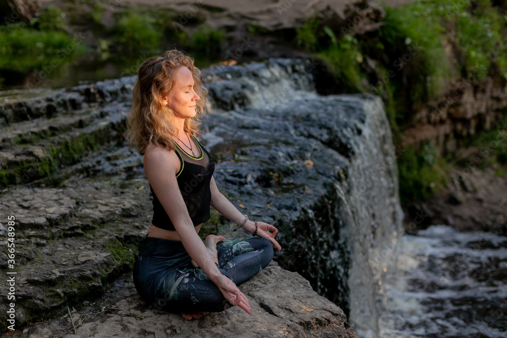 Young woman practicing breathing yoga outdoors in harmony with nature. Fitness girl doing a stretching exercise and meditating on the edge of a waterfall on a forest river.