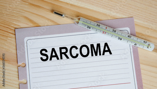 On the sheet is the inscription SARCOMA, next to the thermometer, against the background of a wooden table. A medical concept. photo