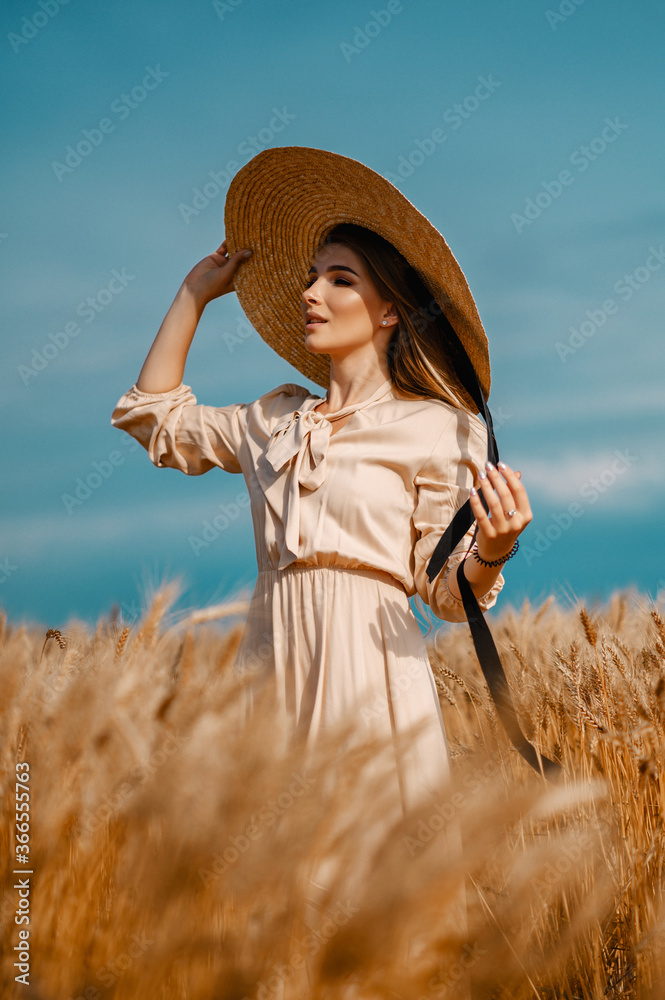 Obraz premium Attractive and beautiful girl in a beige midi dress with a big straw hat, in a wheat field